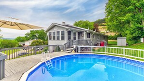 A residential swimming pool with a wooden deck, a covered patio area, and a house in the background.