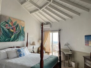 Une chambre à coucher avec un lit à baldaquin, un tableau représentant des fleurs tropicales, un bureau et une vue par la fenêtre.