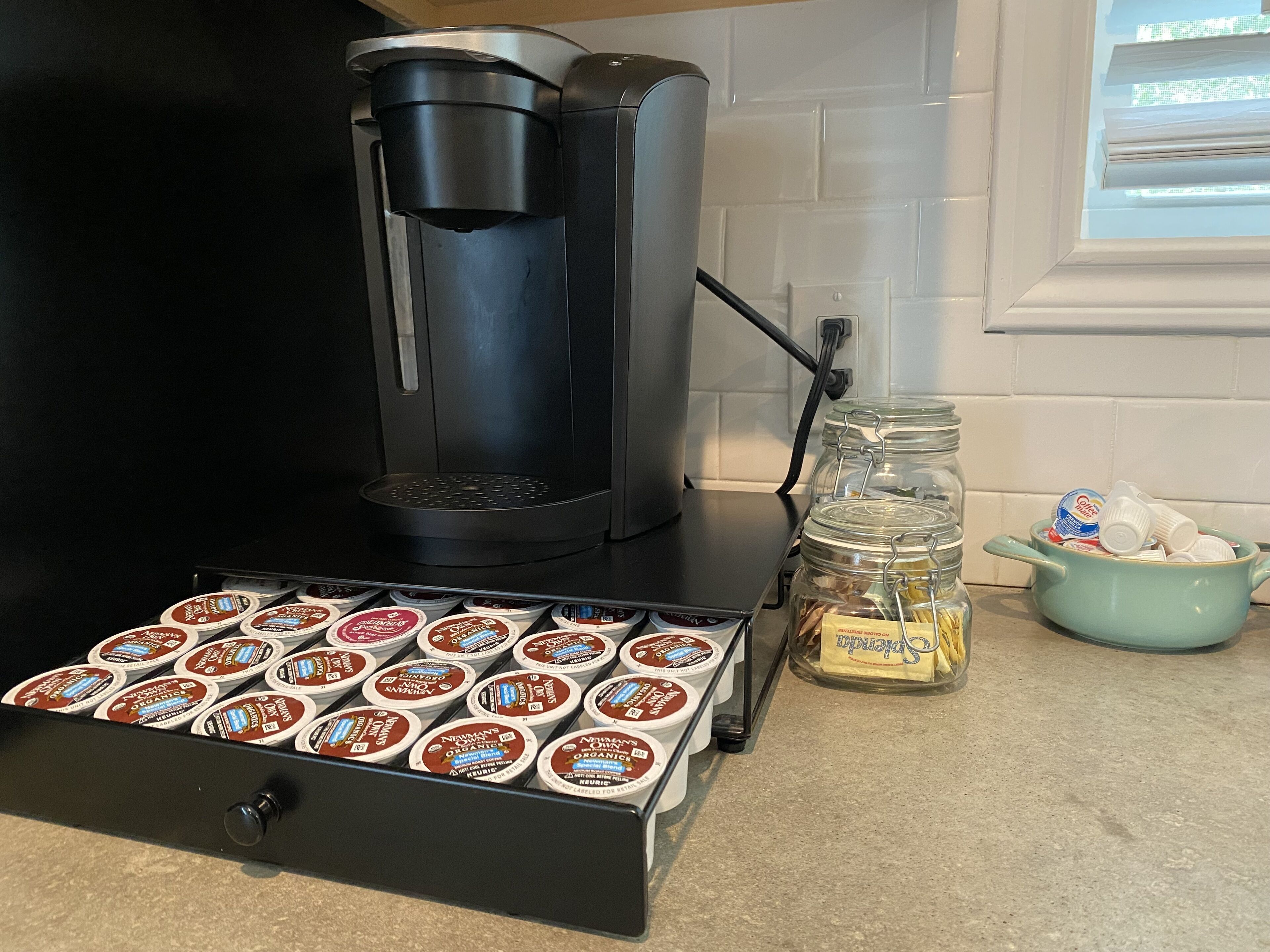 A coffee maker on a kitchen counter with a drawer of coffee pods and a jar of butter.