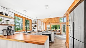A modern kitchen with wooden cabinets, stainless steel appliances, and large windows offering a view of the outdoors.