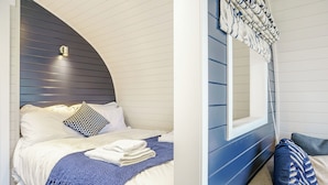 A neatly made bed with white linens and a blue knitted throw, a patterned pillow, and a window with a curtain.