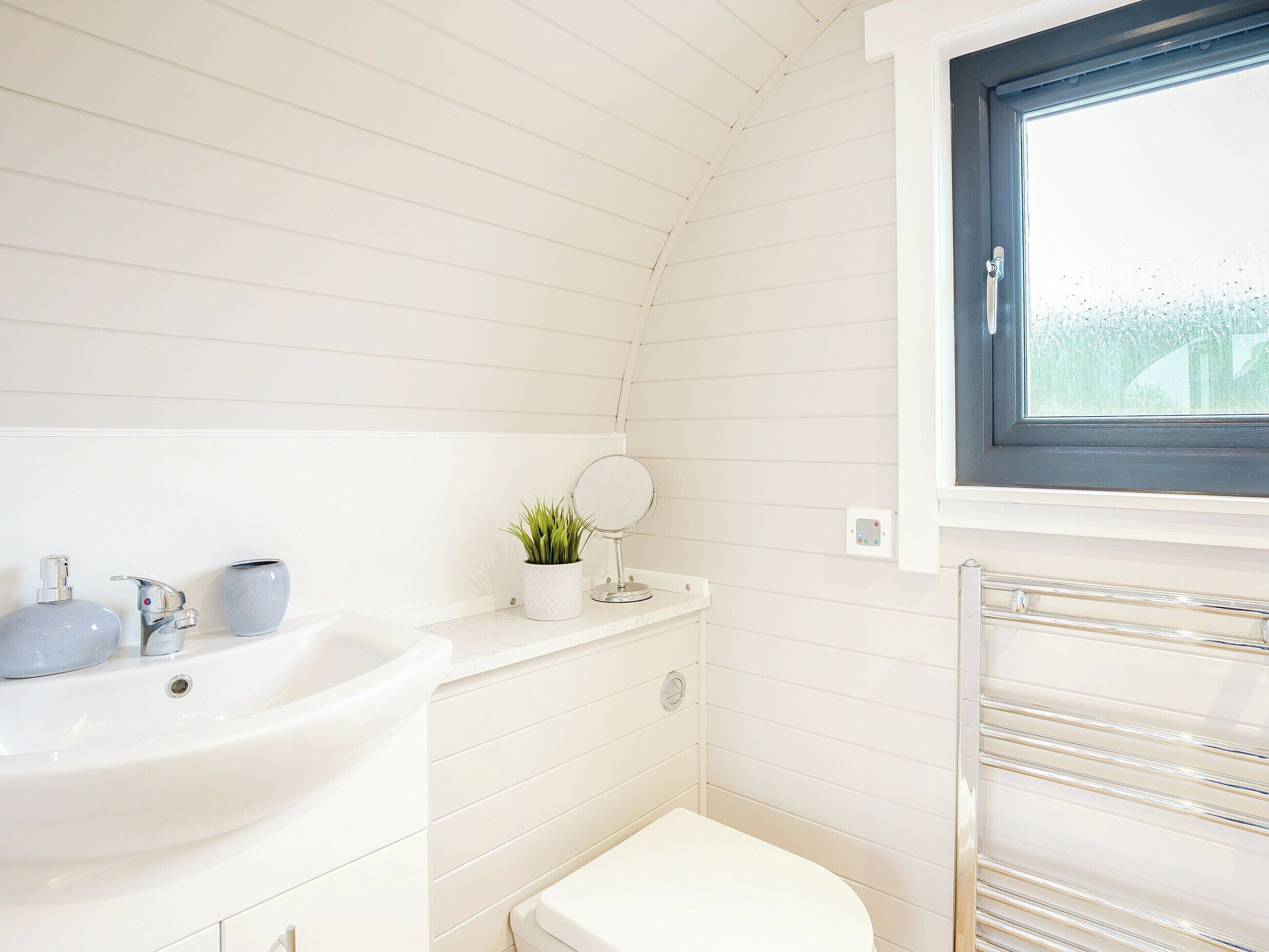 A bathroom with a white sink, a toilet, a small potted plant, a round mirror, and a window with a view of trees.