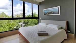 Une chambre avec un grand lit, un ventilateur de plafond, une vue sur la verdure et un tableau encadré au mur.