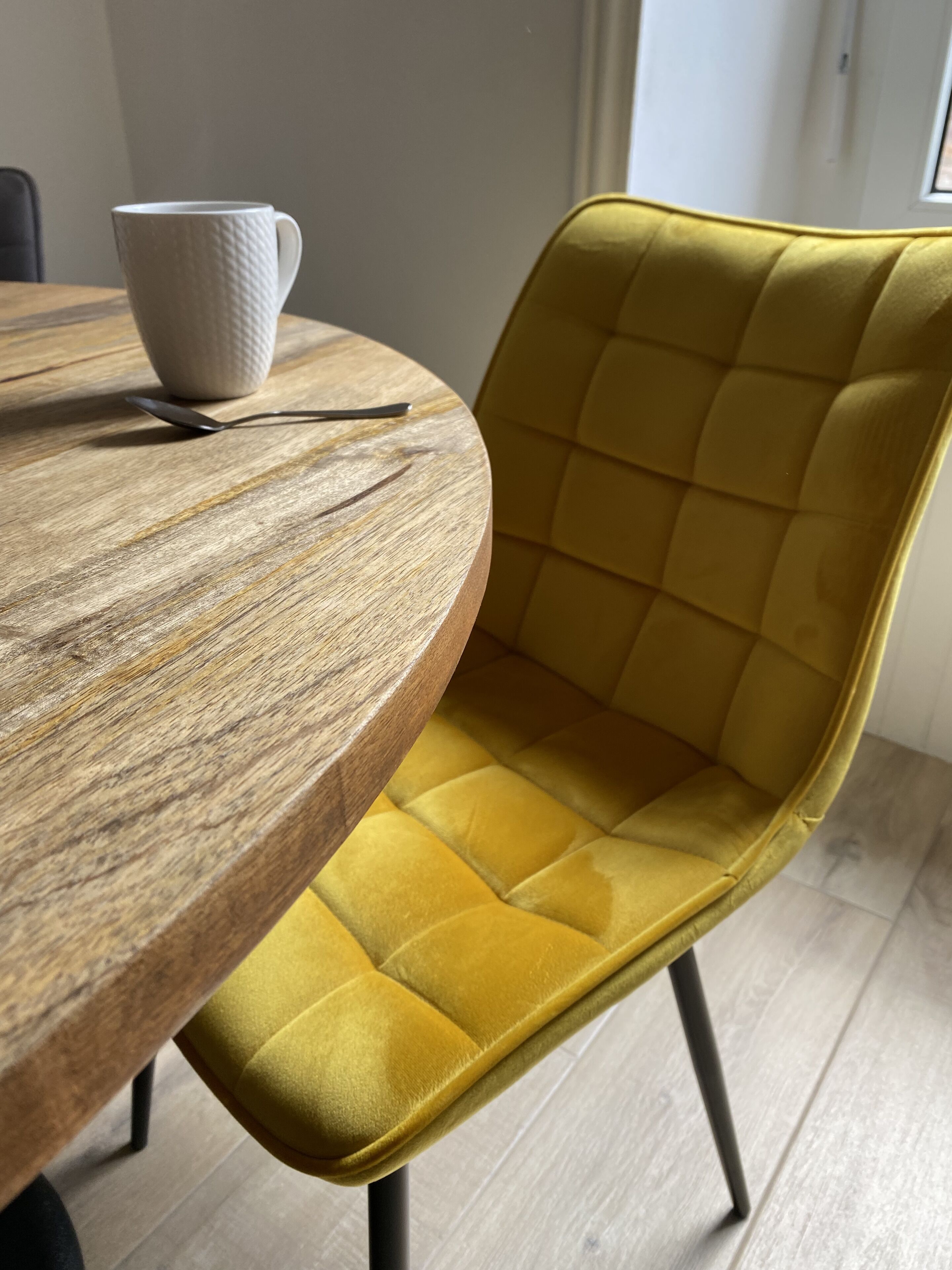 A round wooden table with a white textured mug and a spoon. A mustard yellow upholstered chair with a tufted backrest is positioned next to the table.