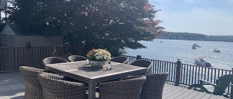 A wooden deck with wicker furniture and a table with a floral centerpiece, overlooking a calm body of water with boats.