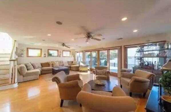 A spacious living room with a large sofa, armchairs, and a coffee table. There are ceiling fans and large windows providing natural light.