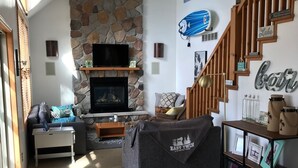 A cozy living room with a stone fireplace, a television, and a staircase leading to another level. - East Twin Lake House - 2 Minutes to town (Lewiston)