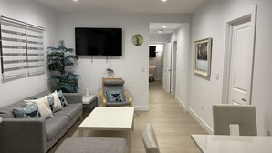 Una sala de estar moderna con un sofá gris, una mesa de centro blanca y un reposapiés gris. Hay un televisor de pantalla plana montado en la pared, una planta en maceta y una silla de madera con cojín.