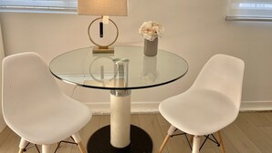 Una mesa redonda de vidrio con dos sillas blancas, una lámpara y un florero con flores.