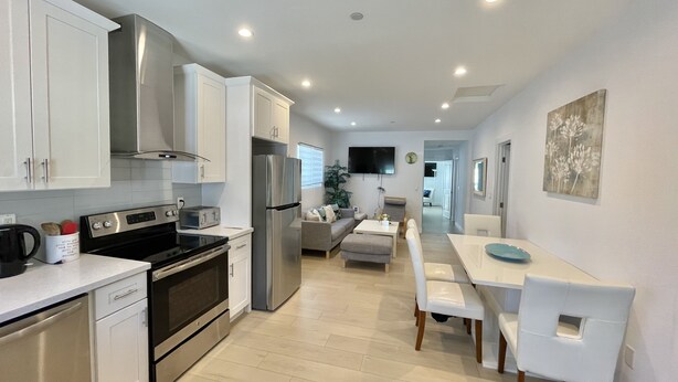 Una cocina moderna con gabinetes blancos, electrodomésticos de acero inoxidable y un comedor con sillas y mesa blancos.