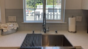 Una cocina con un lavamanos de acero inoxidable, una ventana que ofrece vistas a coches aparcados y árboles, y un rollo de toallas de papel sobre la encimera.