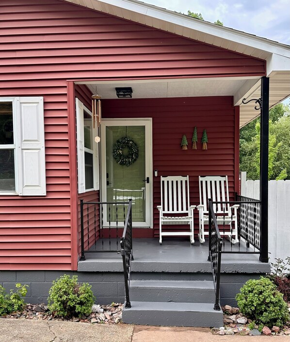 A red house with a porch, a door with a wreath, two white chairs, and a small garden.