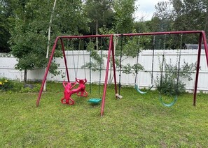 A red swing set in a grassy yard with a white fence and trees in the background. - Charming Red House • Private Yard • Fire Pit (Marquette)