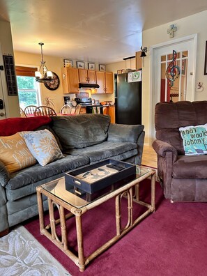 A living room with a grey sofa, a brown armchair, a glass coffee table, and a kitchen in the background.