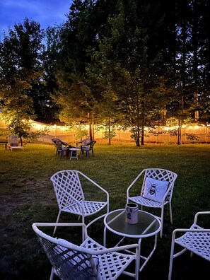 A patio area with white metal chairs and tables, surrounded by trees and a wooden fence. - Charming Red House • Private Yard • Fire Pit (Marquette)