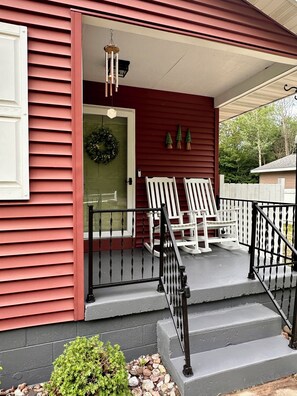 A porch with two white rocking chairs, a black metal railing, and a small potted plant. - Charming Red House • Private Yard • Fire Pit (Marquette)