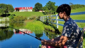 Una persona pesca in un lago tranquillo con una recinzione bianca e una casa con tetto rosso sullo sfondo.