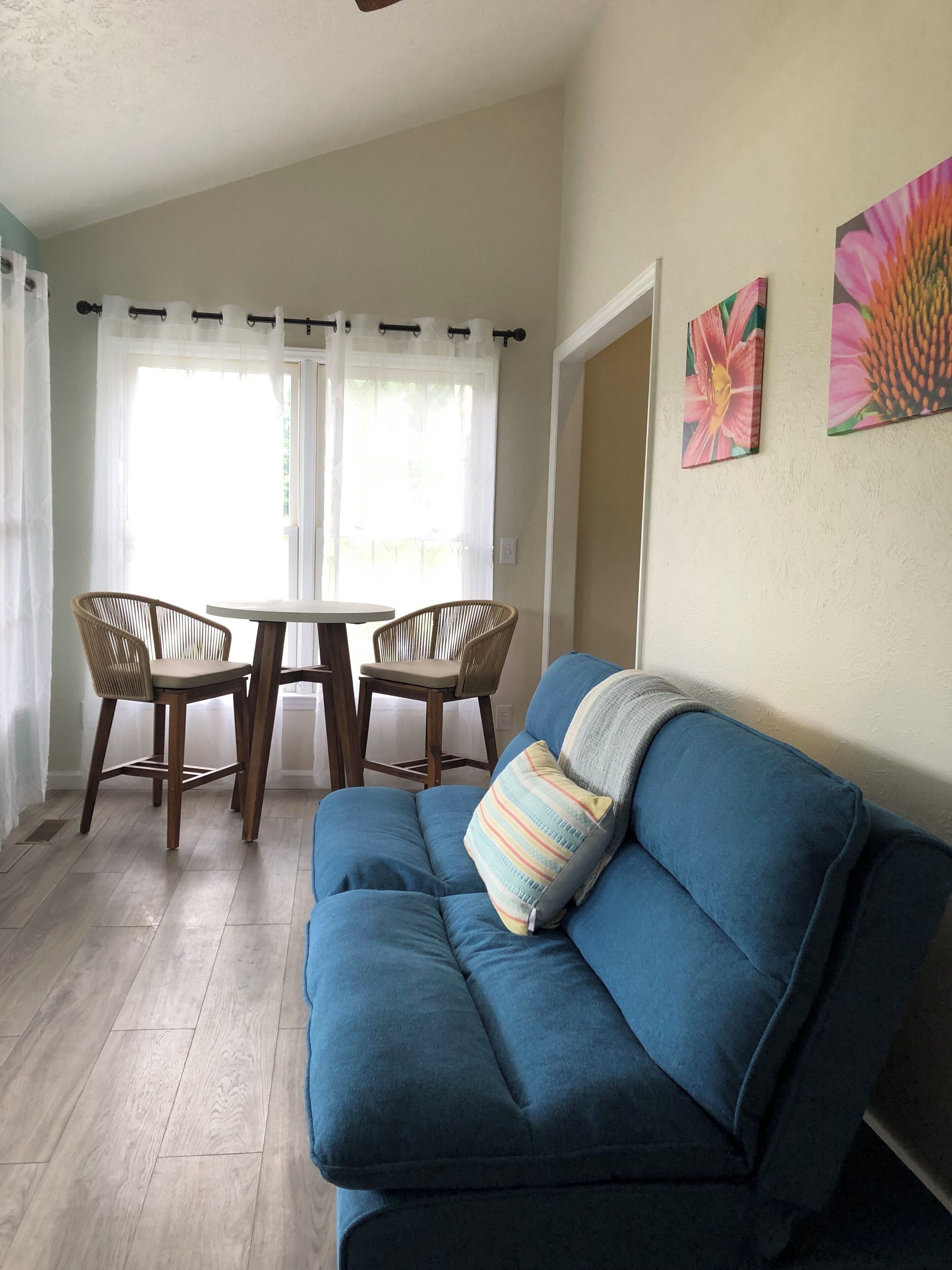 Una sala de estar con un sofá azul, dos sillas de mimbre, una mesita y una ventana con cortinas translúcidas.