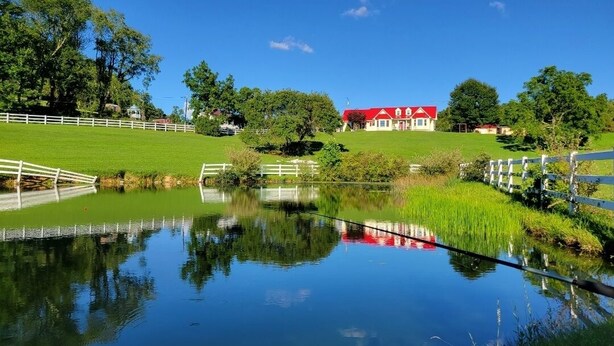 Una tranquilla scena con un lago, un edificio dal tetto rosso, recinzioni bianche e una vegetazione rigogliosa.