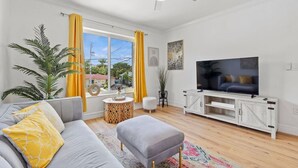 Una sala de estar moderna con un sofá gris, cojines amarillos, un televisor de pantalla plana sobre un mueble blanco, un ventilador de techo y un ventanal con cortinas amarillas.