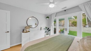 Une chambre à coucher comprenant un lit, une commode, un miroir et donnant sur un jardin grâce à une porte-fenêtre coulissante.