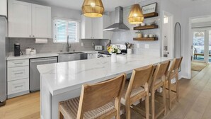 Une cuisine moderne avec des meubles blancs, un grand îlot central doté de tabourets, des luminaires suspendus et une vue sur l’extérieur.
