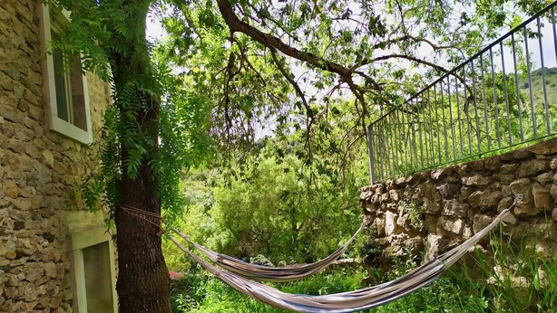 Un hamac est tendu entre deux arbres dans un jardin luxuriant, bordé de murs de pierre et abritant un bâtiment rustique.