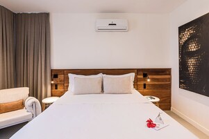 Habitación de hotel con una cama grande, cabecera de madera, unidad de aire acondicionado blanca, sofá blanco, una mesita redonda y un cuadro abstracto de gran tamaño en la pared.
