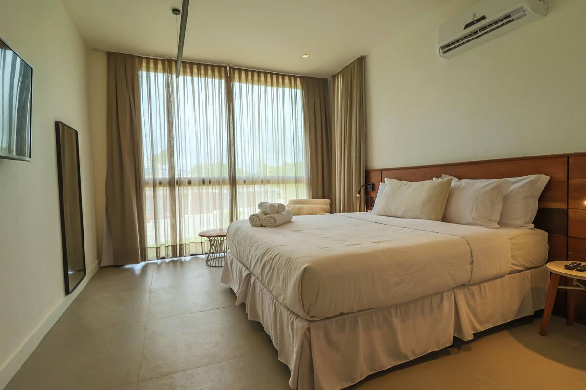 A hotel room with a large bed, a small table, a chair, and a large window with curtains.