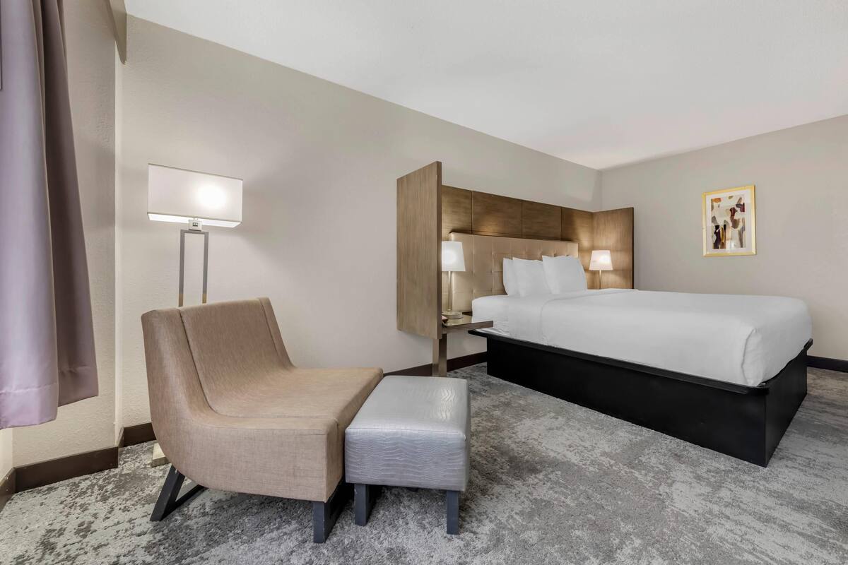 Una habitación de hotel con cama, una silla, una mesita, una lámpara y un cuadro en la pared.