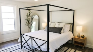 Een hemelbed met een witte spreideken en kussens, een houten nachtkastje met een lamp, en een zwart-wit gestreept vloerkleed.