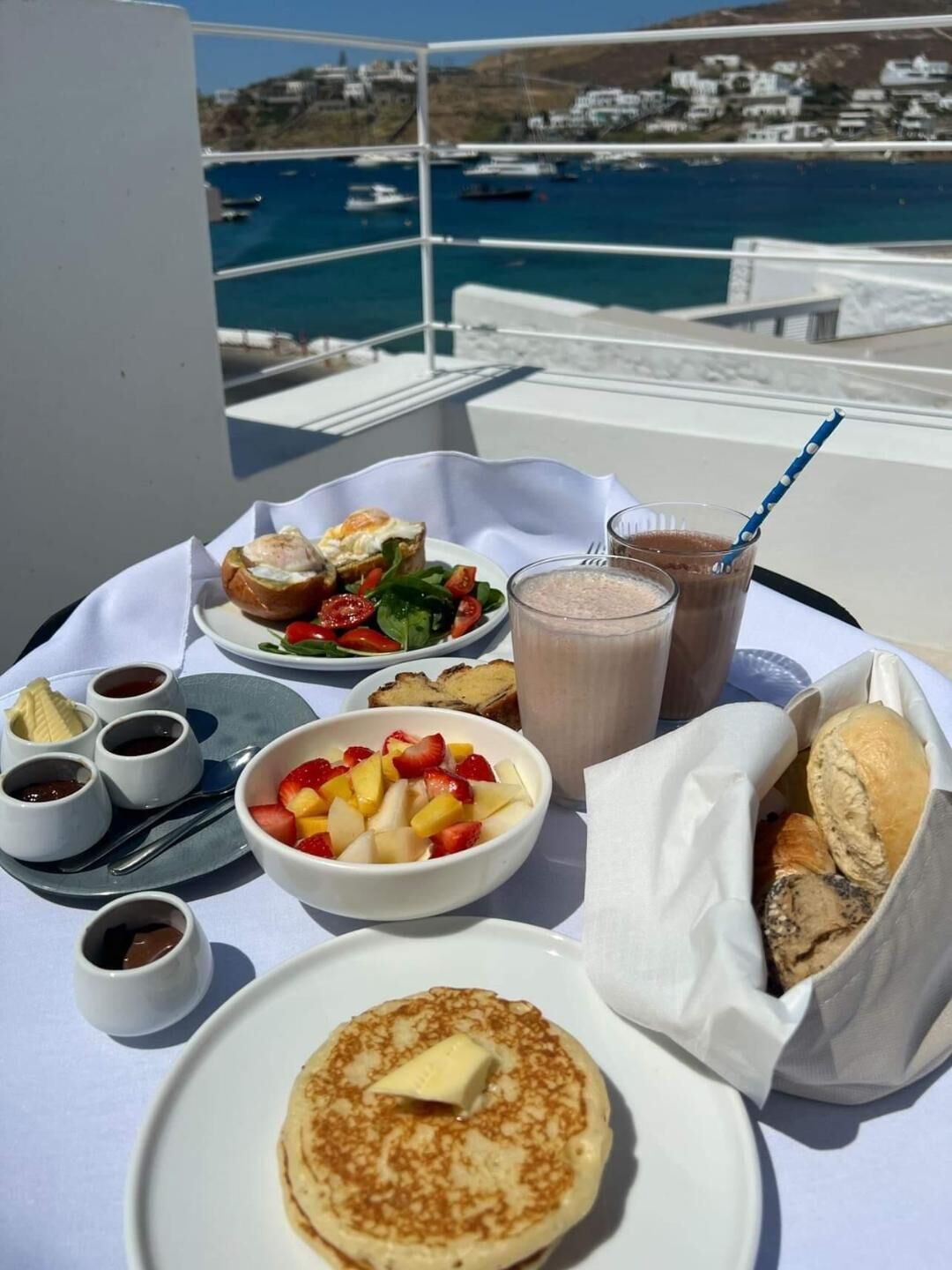 A table with breakfast items including pancakes, fruit, and beverages, with a scenic view of the sea and buildings in the background.