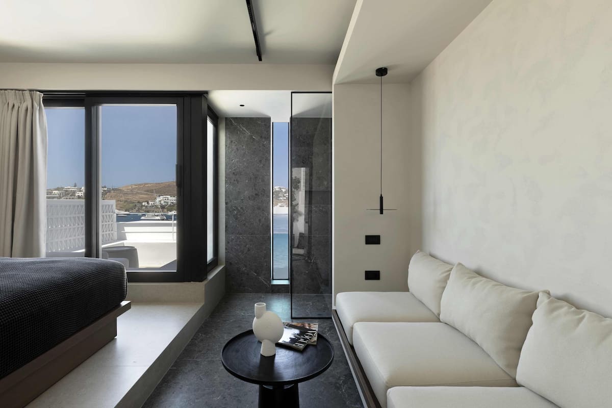 A modern hotel room with a large bed, a white sofa, a small round table with a vase and a magazine, and a view of the city through large windows.