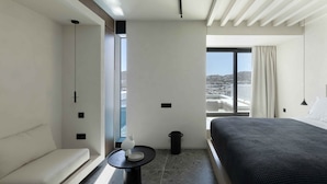 A modern hotel room with a bed, a sofa, a small table, and a view of the city. - CUBIC Mykonos Seafront Design Suites (Mykonos)