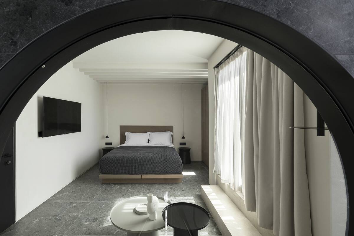 A modern bedroom with a bed, bedside tables, a round mirror, and a view through an arched doorway.