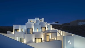 A modern, multi-story building with illuminated windows and balconies, set against a rocky hillside at dusk. - CUBIC Mykonos Seafront Design Suites (Mykonos)