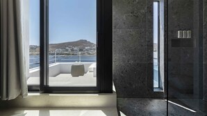 A modern bathroom with a glass shower enclosure and a view of a cityscape and water body. - CUBIC Mykonos Seafront Design Suites (Mykonos)