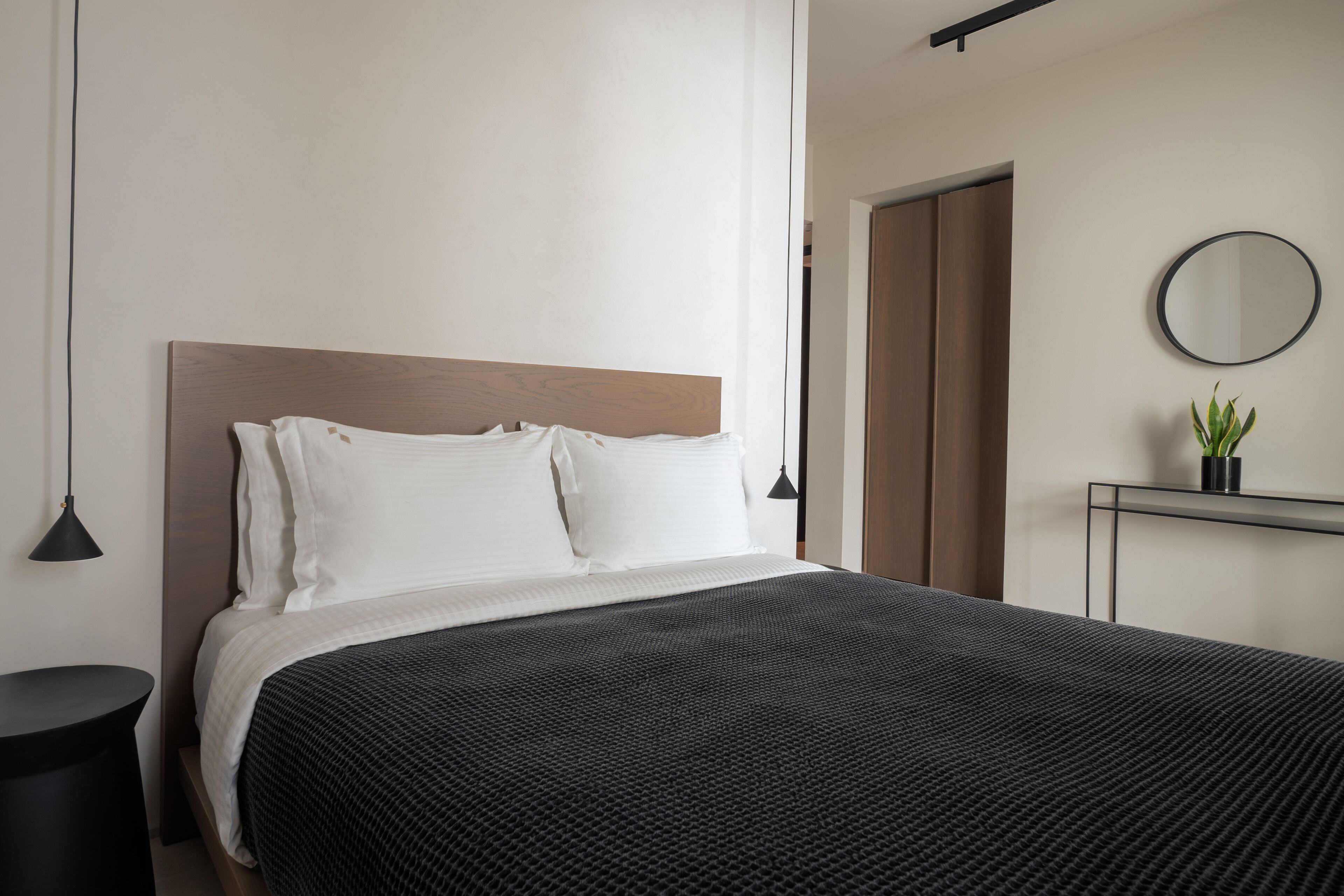 A modern bedroom with a wooden headboard, white bedding, a black textured bedspread, a round mirror, and a small potted plant on a bedside table.