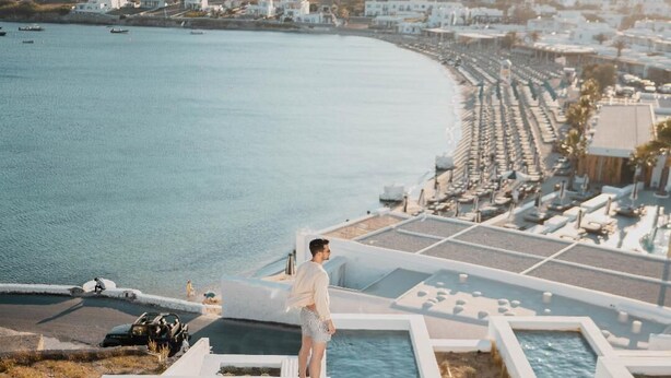 A man standing by a pool with a view of a coastal town and a hill in the background. - CUBIC Mykonos Seafront Design Suites (Mykonos)