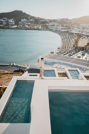 A man standing by a pool with a view of a coastal town and a hill in the background. - CUBIC Mykonos Seafront Design Suites (Mykonos)