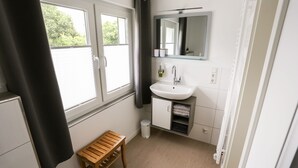 A bathroom with a window, a sink, a mirror, a small wooden stool, and a trash bin. - Moderne Ferienwohnung in Kassel (Kassel)