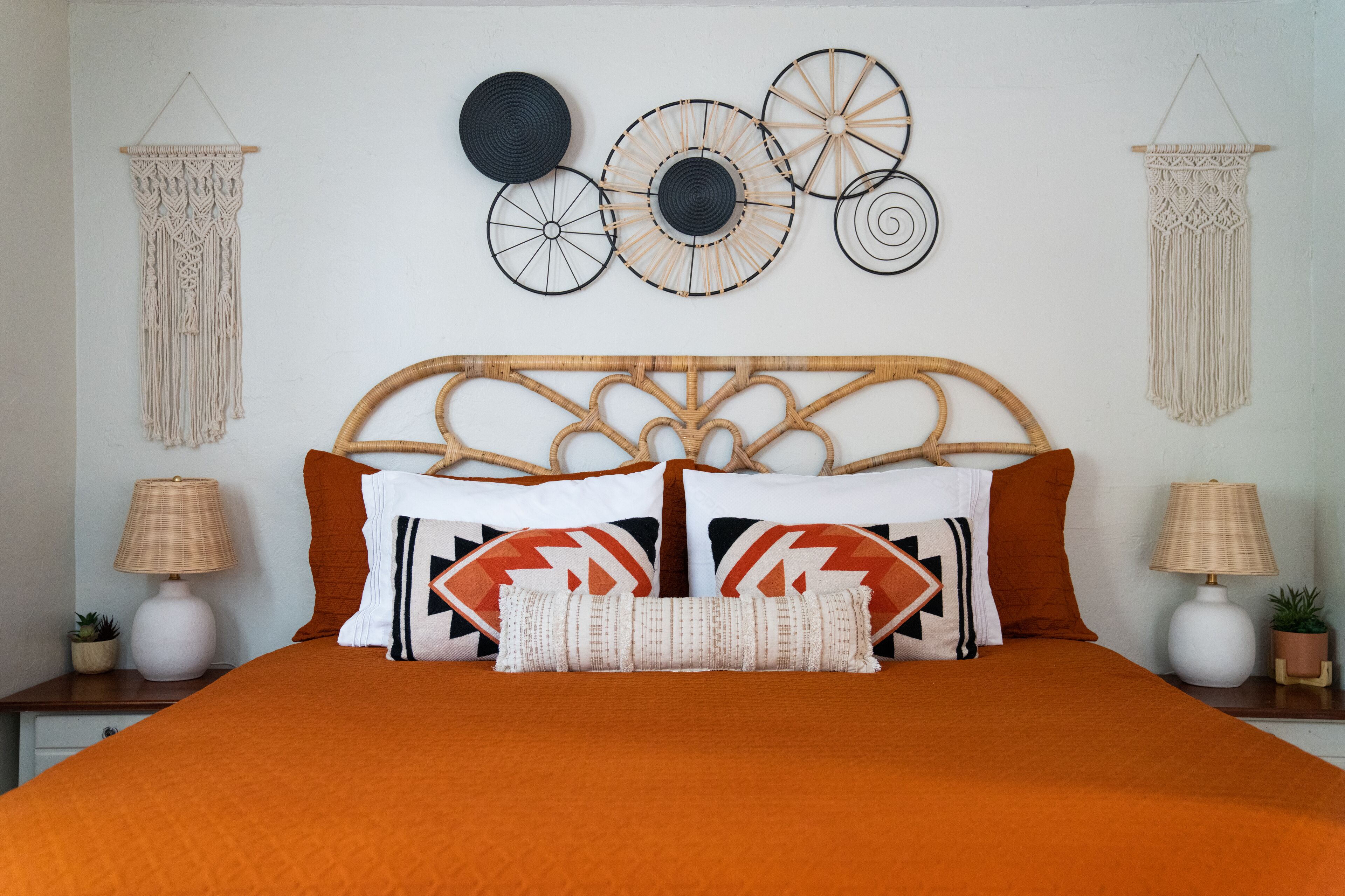 Un letto con una coperta arancione, due cuscini con motivi geometrici e un arazzo macramè appeso al muro.