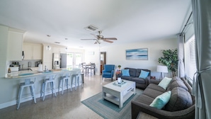 A modern kitchen with white cabinets, a central island with bar stools, a dining area with a table and chairs, a living room with a sofa and cushions, and a ceiling fan.