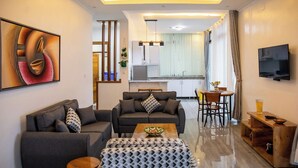 A modern living room with a sofa, ottoman, and coffee table, a dining area with a table and chairs, a kitchen in the background, and a TV mounted on the wall.