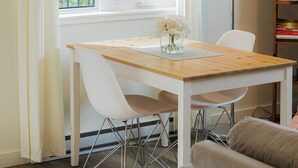 Un comedor con una mesa de madera, sillas blancas, una lámpara de pie y un ventanal con cortinas.