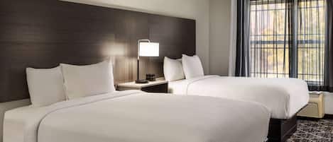 A hotel room with two beds, a bedside table with a lamp, and a large window with curtains.