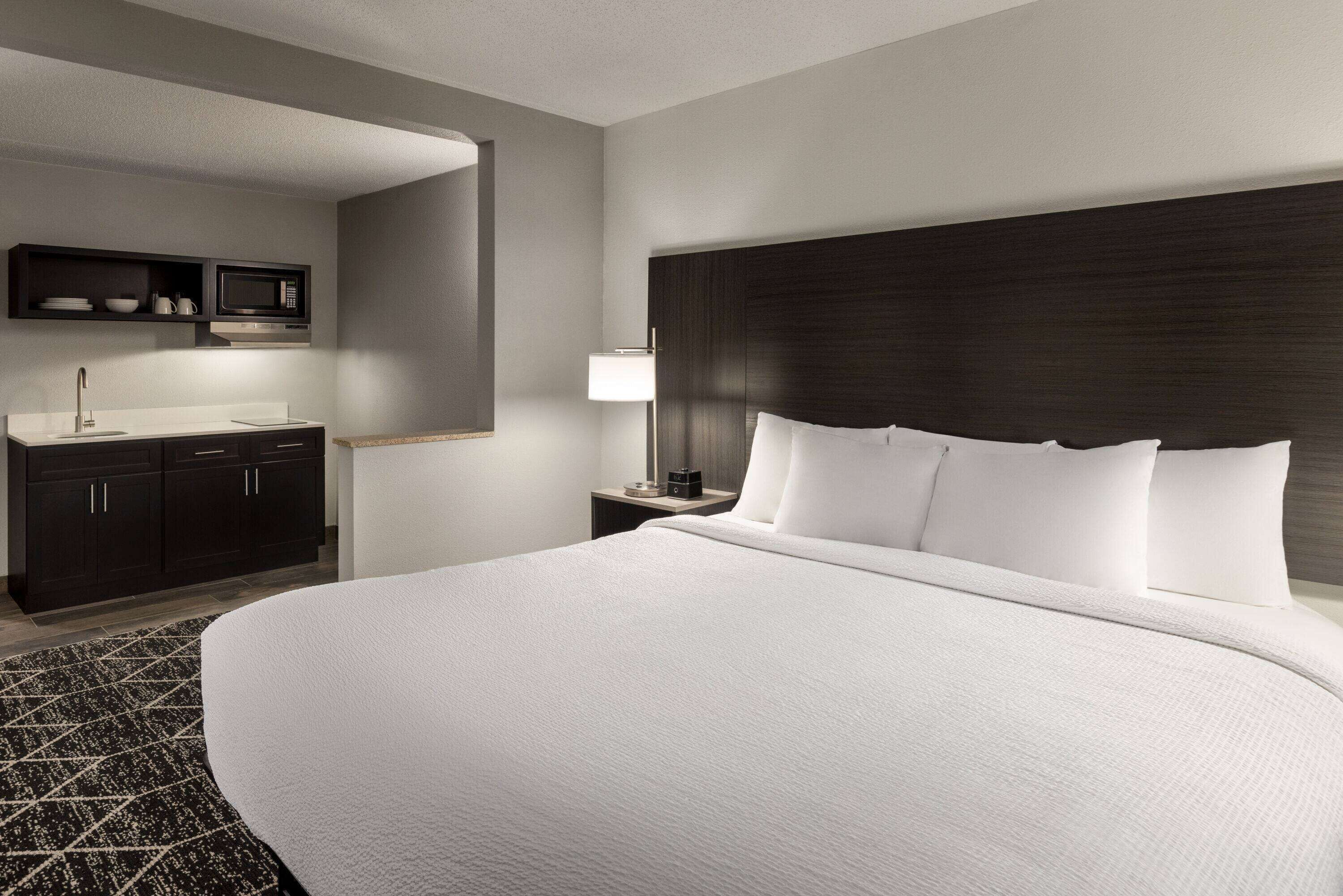 Une chambre d’hôtel avec un grand lit, une tête de lit en bois foncé, une table de chevet avec une lampe, une petite étagère avec un micro-ondes et une armoire en bois foncé.
