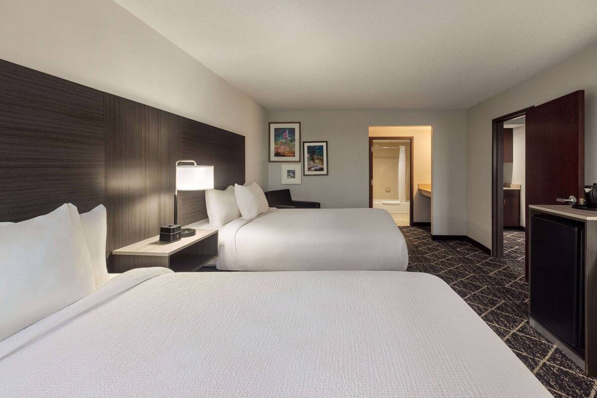 Ein Hotelzimmer mit zwei Betten, einem Schreibtisch mit Lampe, einem Kleiderschrank und gerahmten Bildern an der Wand.