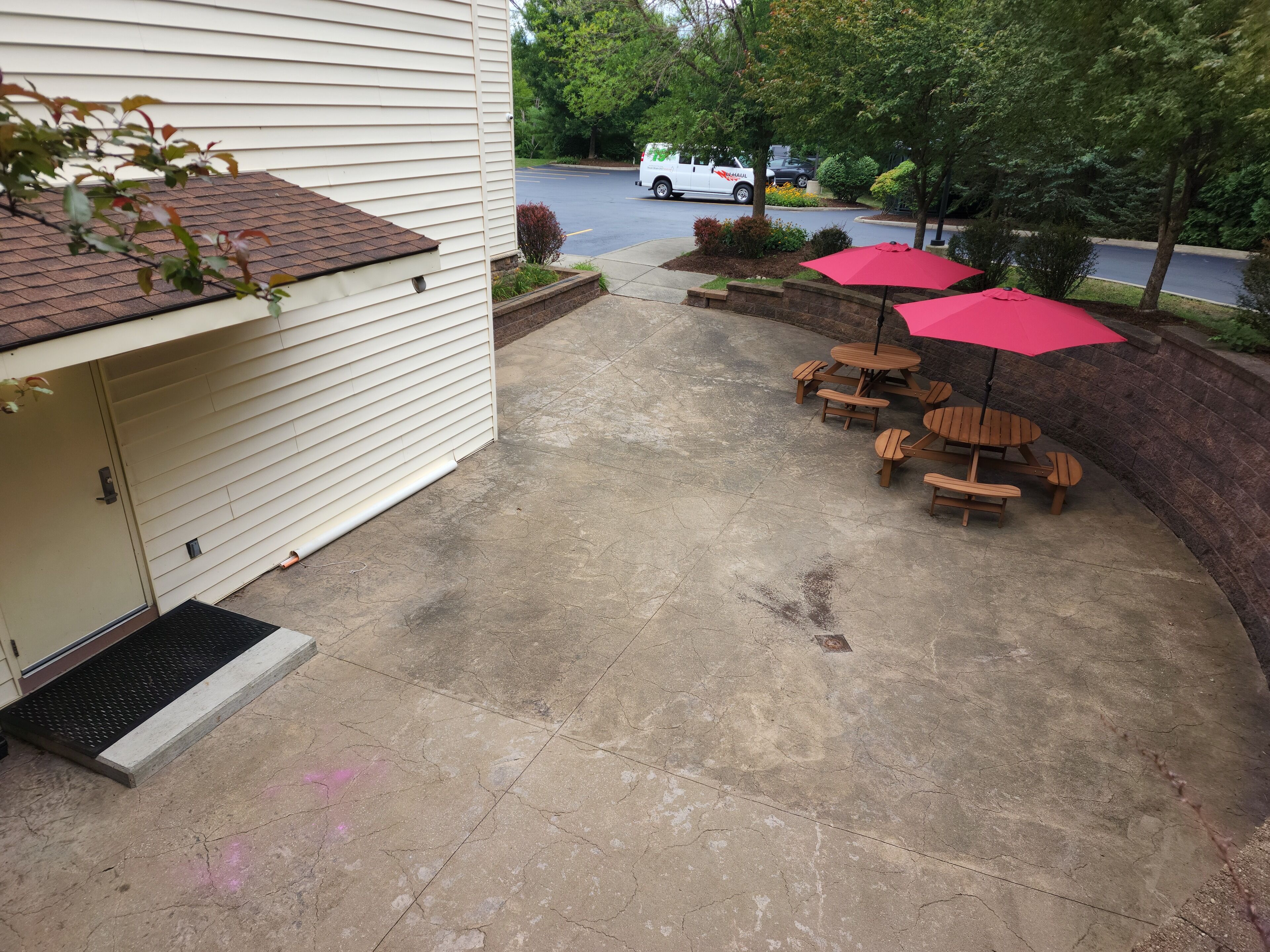 A patio with a concrete floor, wooden tables and chairs, and a red umbrella.