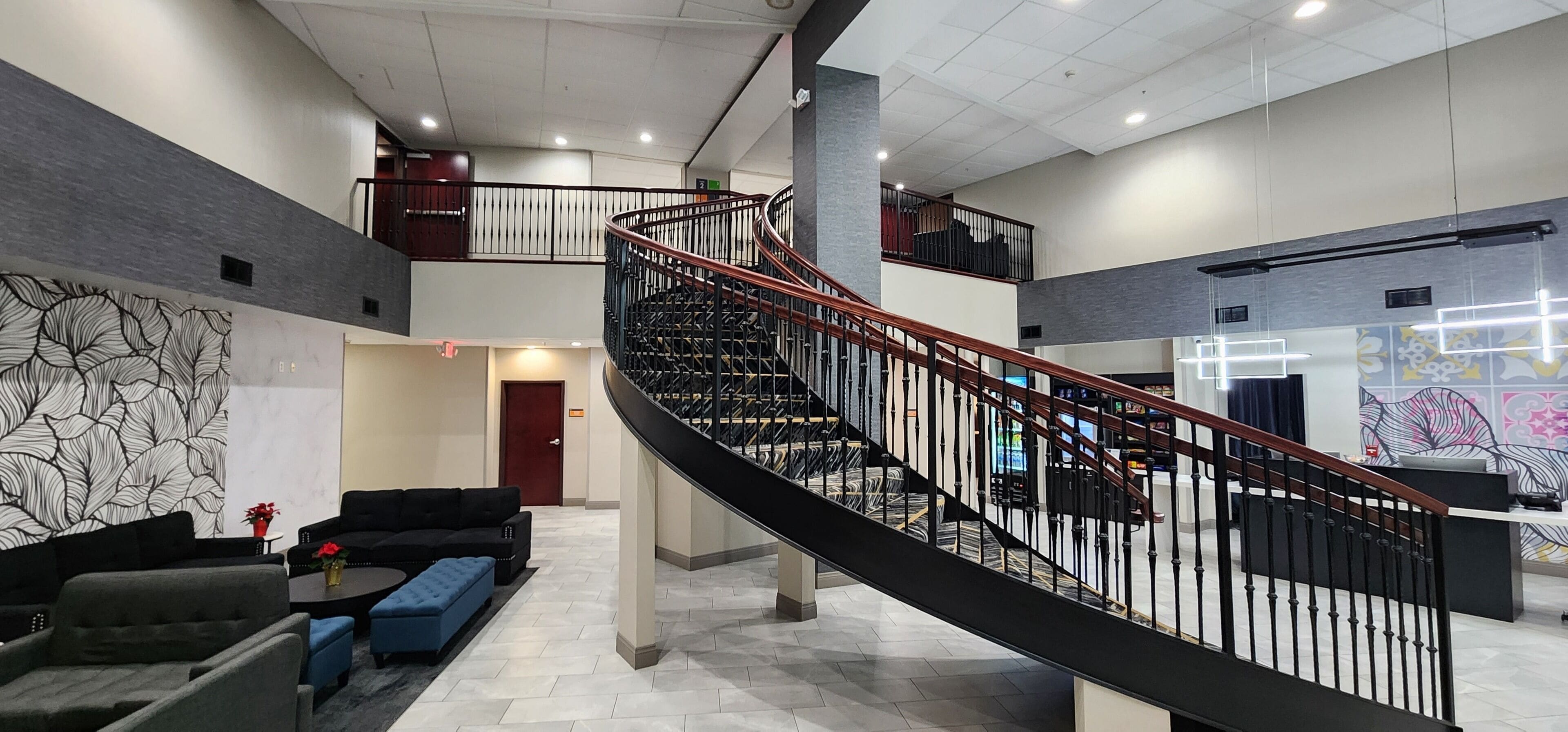 A spacious lobby with a curved staircase, a black leather sofa, a blue ottoman, and a coffee table with a vase of red flowers.